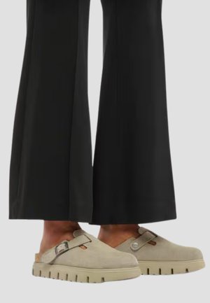 Scarpe slip-on beige chiaro con cinturini con fibbia e suole spesse, indossate con pantaloni neri a gamba larga.