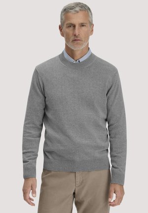 Mann mit grauen Haaren und Bart, der einen grauen Rundhalspullover über einem hellblauen Hemd mit Kragen und beigefarbenen Hosen trägt, steht vor einem einfarbigen Hintergrund.