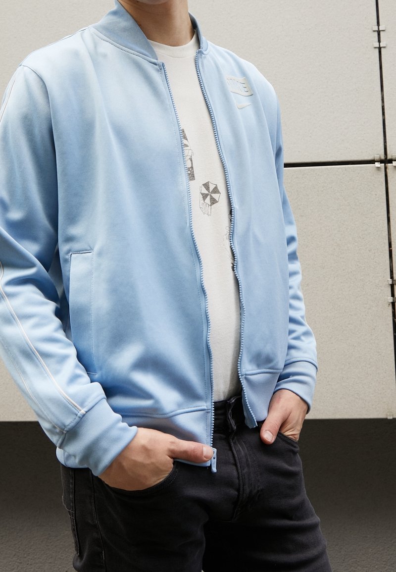 Giacca bomber azzurro chiaro con una texture liscia, zip completa, tasche inclinate e dettagli a strisce bianche sulle maniche, indossata sopra una t-shirt con grafica.