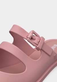 Rosafarbener Riemen einer Gummisandale mit verstellbarer Schnalle und eingeprägtem Markennamen "Melissa" auf einfachem Hintergrund.