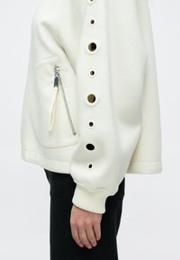 Λευκό φλις φουτερ με κυκλικές εγκοπές κατά μήκος του μανικιού, με πλαϊνή τσέπη με φερμουάρ και ριμπ μανσέτες για επιπλέον υφή.