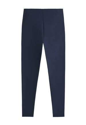 Femme debout portant une veste polaire zippée rayée noire et blanche, des leggings bleu marine, des chaussettes blanches et des baskets beige sur fond gris.