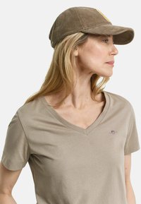 Beige V-hals T-shirt i blødt stof med korte ærmer, der har et lille broderet design på brystet og en brun kordfløjl cap.