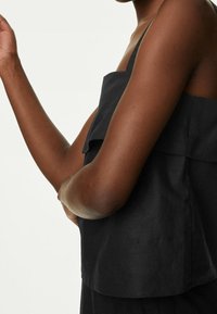 Camisole en lin noir avec un design à couches, fines bretelles et une coupe ample, présenté sur un fond neutre.