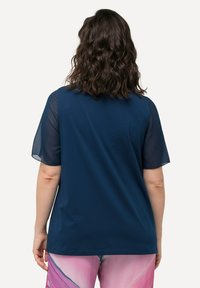 Námořnická modrá tričko s průhlednými krátkými rukávy, volný střih a hladká textura. Viditelné švy podél lemu, nošeno s vzorovanými růžovými kalhotami.