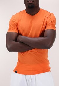 Oranžové tričko s krátkým rukávem z měkké látky s kulatým výstřihem. Obsahuje malou černou nášivku na lemu. Přiléhavý design zvýrazňuje torzo.