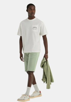 T-shirt bianco a maniche corte con tasca, pantaloni corti verde chiaro e sneakers beige; tiene una camicia verde. Design semplice, vestibilità casual.