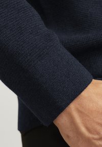 Marinerenblauwe gebreide trui-mouwen met een textuurrijke, geribbelde patroon. De stof lijkt zacht met een licht losse fit, wat het comfort benadrukt.