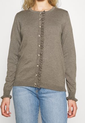 Kobieta nosząca brązowy sweter na guziki z falbanką z przodu i na mankietach, w połączeniu z jasnoniebieskimi dżinsami.