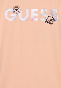 T-shirt w kolorze brzoskwiniowym z dużym haftowanym napisem "GUESS". Posiada okrągłą grafikę z tekstem i sercem, wykonana z materiału bawełnianego.