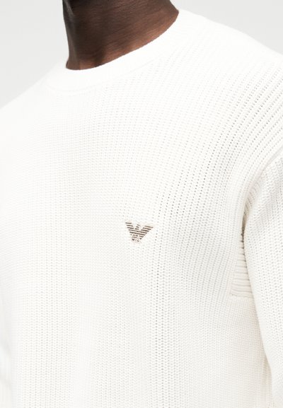 Witte gebreide trui met een gestructureerd patroon, voorzien van een klein beige logo op de borst. Ronde halslijn en geribbelde manchetten.