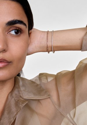 Due due bracciali delicati, uno in oro rosa e uno in argento, adornati con piccole pietre chiare, indossati su un polso, abbinati a una blusa beige trasparente.