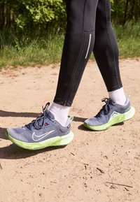 Osoba w czarnych legginsach, białych skarpetach i niebiesko-zielonych butach do biegania Nike stoi na nasłonecznionej, piaszczystej ścieżce w pobliżu zielonego listowia.