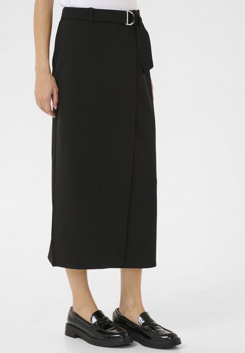 Jupe midi noire avec ceinture et boucle argentée, portée avec des mocassins noirs brillants, montrée de la taille jusqu’aux pieds sur fond blanc.