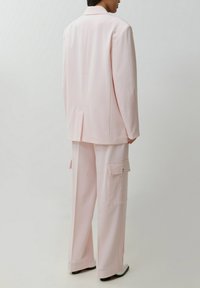 Personne portant un blazer oversize rose pâle et un pantalon cargo large assorti, debout devant un fond blanc uni.