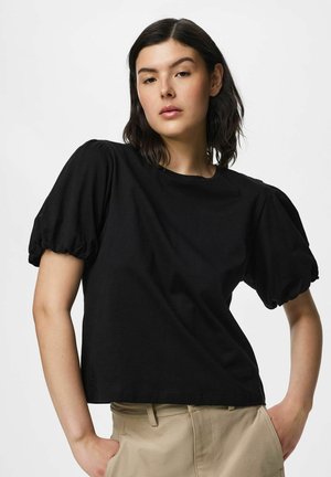 Jonge vrouw met donker haar die een zwarte top met pofmouwen en beige broek draagt, poserend met handen in de zakken tegen een effen achtergrond.