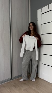 Bílá zkrácená košile s dlouhými rukávy drží osoba, má kulatý výstřih a hladkou látku. Viditelné jsou šedé tepláky a světlá podlaha.