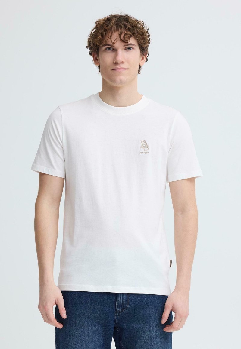 Νέος άνδρας με σγουρά μαλλιά που φοράει λευκό μπλουζάκι με μικρό σχέδιο καρέκλας παραλίας στο στήθος και μπλε τζιν, στέκεται μπροστά σε ένα απλό φόντο.