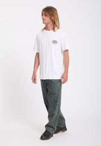 Homme aux cheveux mi-longs portant un t-shirt blanc avec un motif œil et soleil, jean noir délavé et chaussures noires, debout et regardant vers la gauche.
