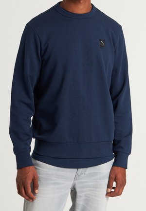 Man wearing navy blue long-sleeve crew neck sweatshirt with small black square logo on chest, paired with light gray jeans.