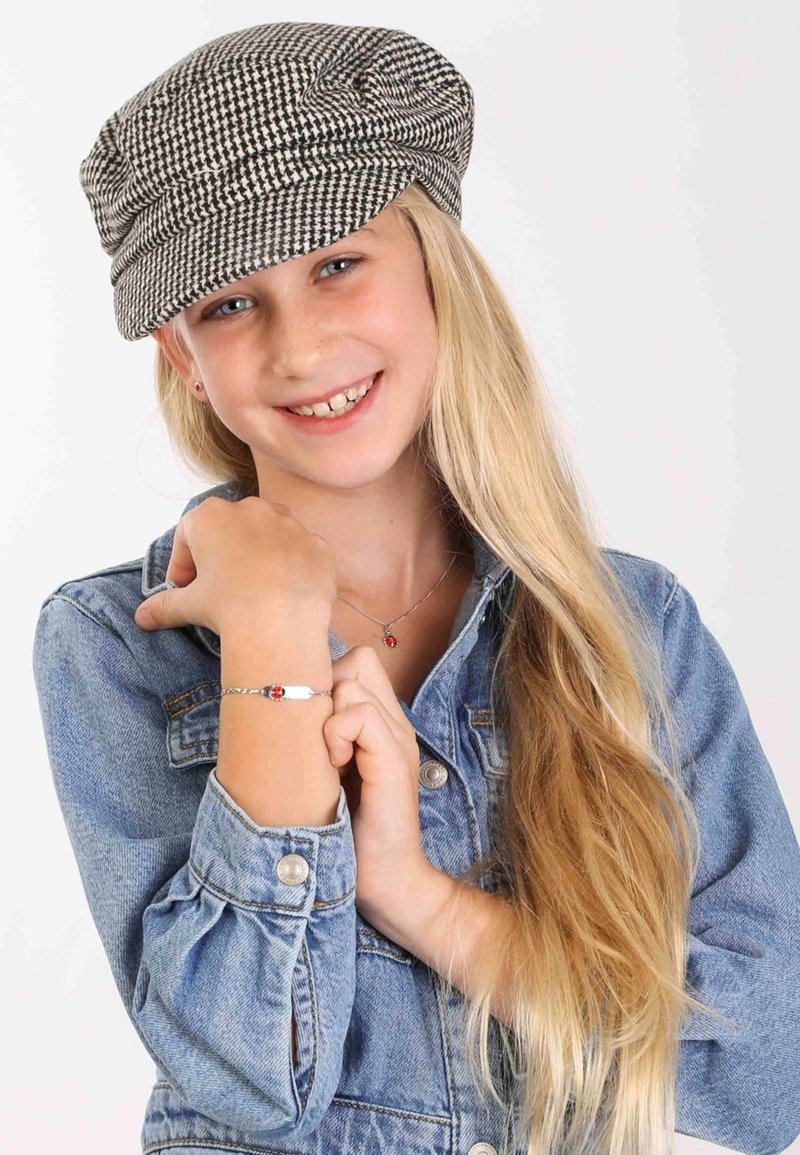 Zwart-wit houndstooth patroon pet, knoopje denimjasje, zilveren armband met rode details, lang blond haar, glimlachende uitdrukking.