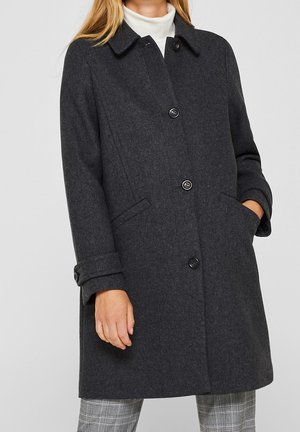 Femme portant un manteau en laine gris foncé boutonné avec col, par-dessus un col roulé blanc et un pantalon à carreaux gris, une main dans la poche.