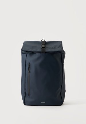 Zaino blu scuro con chiusura a rotolo, tasca frontale con cerniera verticale e fibbia nera su sfondo chiaro uniforme.