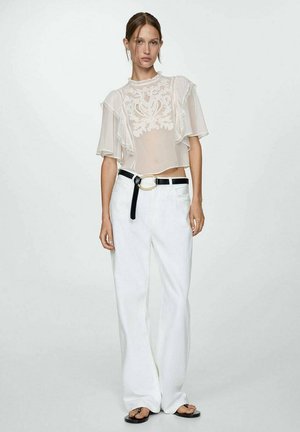 Bílá průsvitná blůza s květinovou výšivkou, s volánkovými rukávy, v kombinaci s širokými bílými kalhotami a černým opaskem se zlatou kulatou sponou.