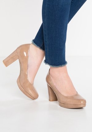 Beige Lackleder-Pumps mit abgerundetem Zehenbereich, ausgestattet mit einer Plateau-Sohle und einem klobigen Absatz, kombiniert mit dunklen Jeans.
