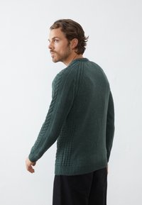 Strikkegenser i mørkegrønn, med kabelmønster på ermene og sidene, ribbestrikkede mansjetter og kant, samt en rund halsdesign.