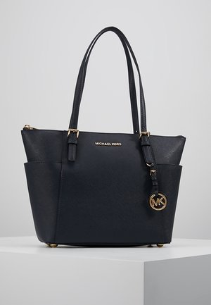 Czarna torba typu tote marki Michael Kors z podwójnymi uchwytami, złotą tabliczką z logo i okrągłym zawieszka MK, prezentowana na białym piedestale na szarym tle.