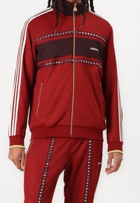 Röd zip-up träningsjacka med tre vita ränder på ärmarna, sammetsliknande brun horisontell panel och dekorativa nitar längs bröstet och sidorna.