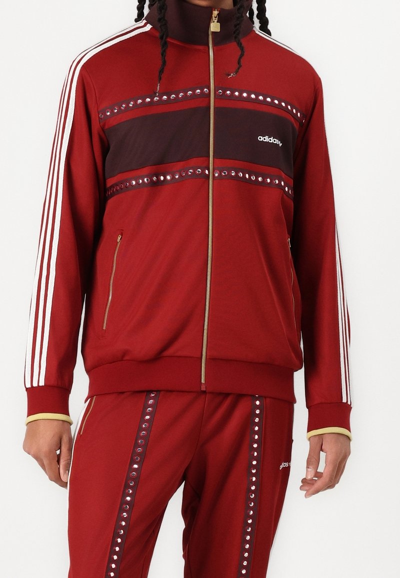 Röd zip-up träningsjacka med tre vita ränder på ärmarna, sammetsliknande brun horisontell panel och dekorativa nitar längs bröstet och sidorna.