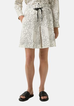 Shorts in cream fabric with black floral outlines, featuring an elastic waistband with a drawstring and side pockets. Worn with black slides.