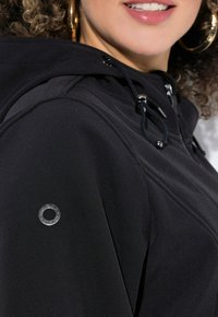 Haut à capuche noir en tissu lisse, avec une fermeture éclair à l'avant, une capuche à cordon de serrage et un accent métallique rond sur la manche.