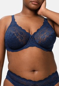 Marinblå spetsbehå med vågig kant, med stöd av bygel och blommönster; parat med matchande spetsunderkläder.