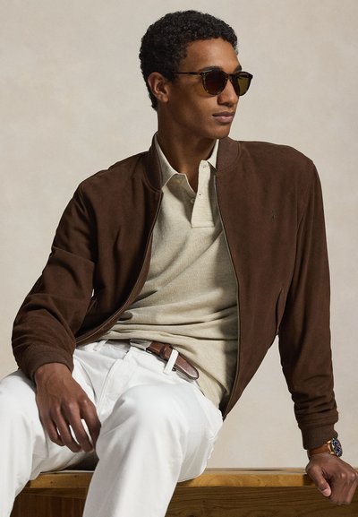 Man met zonnebril, bruine suède jas, beige poloshirt, witte broek en polshorloge, zittend op een houten bank tegen een effen achtergrond.