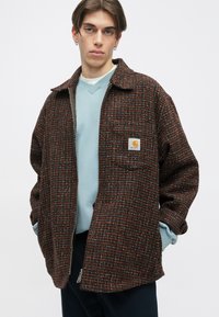 Brun, tekstureret overskjorte med orange, blå og hvide vævede mønstre. Har en lynlåslukning og en brystlomme med et logo-patch.