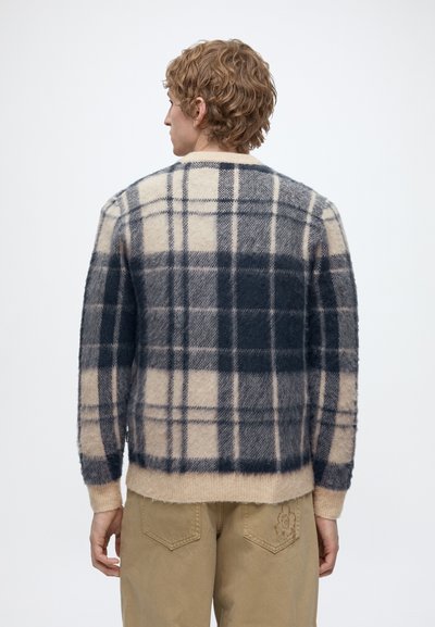 Homme aux cheveux blonds bouclés portant un pull à carreaux beige et gris foncé ainsi qu’un pantalon beige, vu de dos sur un fond uni.