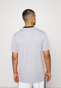 Jersey deportivo gris con cuello negro, acentos naranjas en las mangas, mangas cortas y un ajuste holgado. El material parece ser ligero y transpirable.