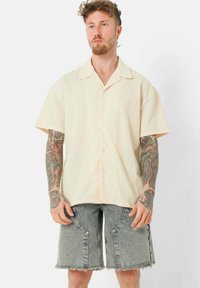Kurzärmliges, hellbeiges Hemd mit strukturiertem Muster, Knopf-Frontdesign und spitzem Kragen, kombiniert mit ausgefransten Jeansshorts.