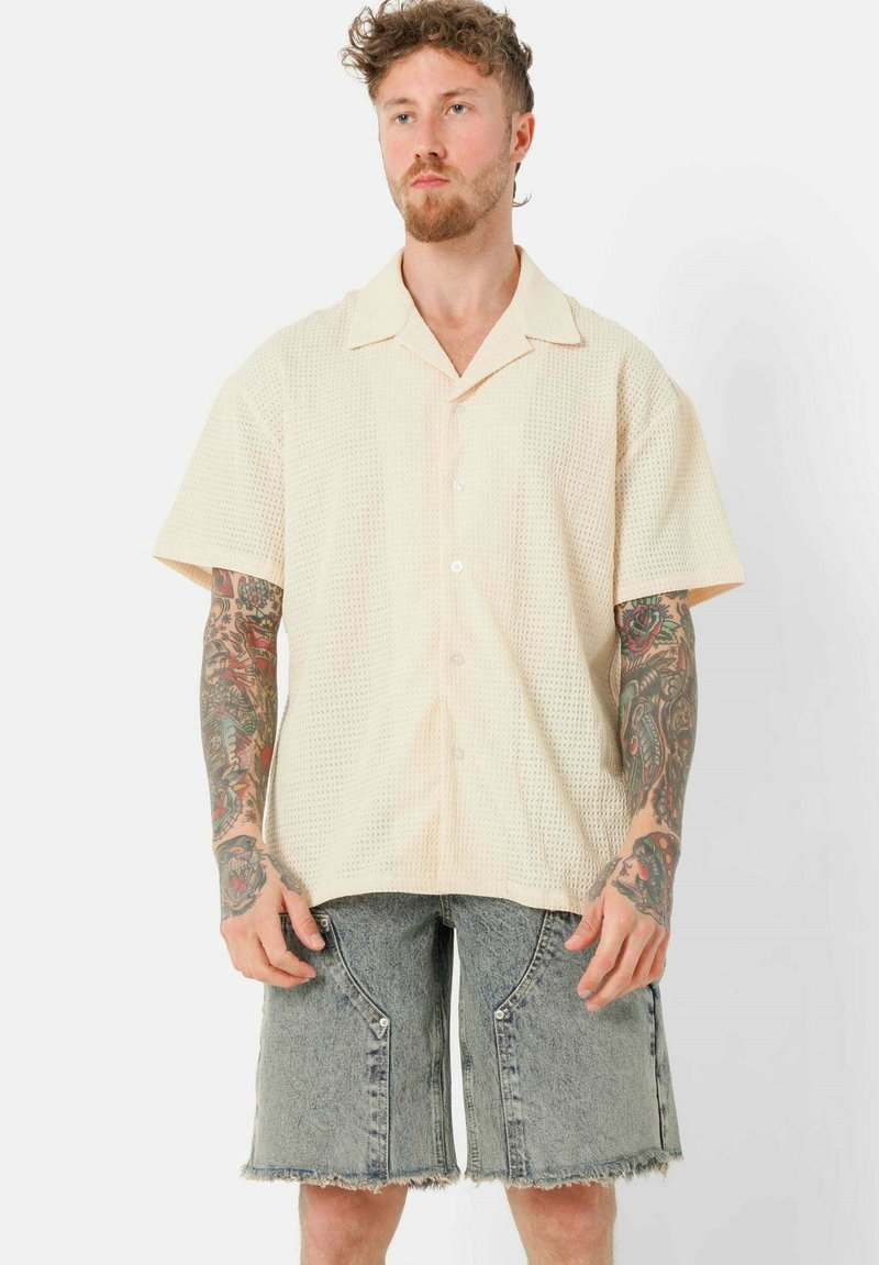Kurzärmliges, hellbeiges Hemd mit strukturiertem Muster, Knopf-Frontdesign und spitzem Kragen, kombiniert mit ausgefransten Jeansshorts.