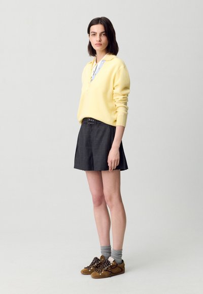 Suéter de punto amarillo, falda plisada gris y zapatillas marrones con cordones decorativos. Calcetines grises visibles por encima de las zapatillas. Diseño sencillo, aspecto moderno.