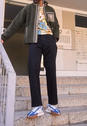 Veste en polaire verte avec des accents gris, t-shirt floral en bleu, jaune et crème, pantalon noir et baskets bleues avec des semelles marron.