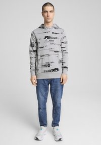 Gray hoodie with black graphic patterns and logos, paired with blue jeans and white sneakers, featuring a relaxed fit and front pocket.