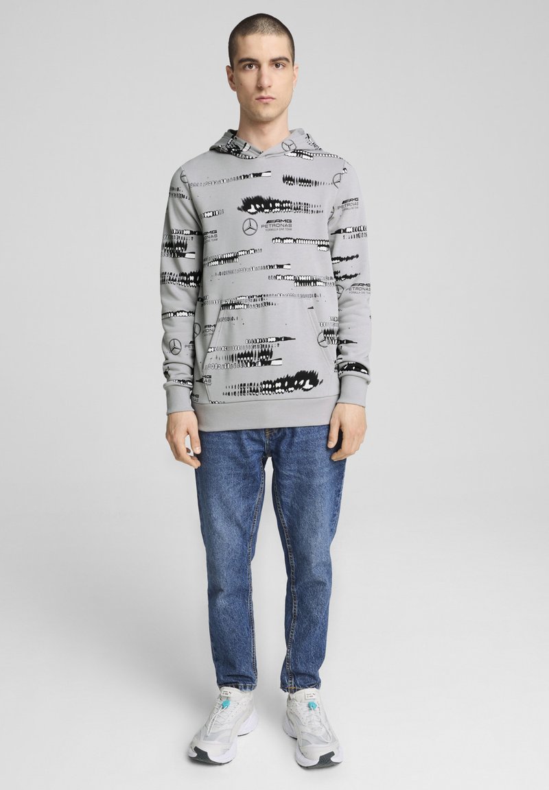 Gray hoodie with black graphic patterns and logos, paired with blue jeans and white sneakers, featuring a relaxed fit and front pocket.