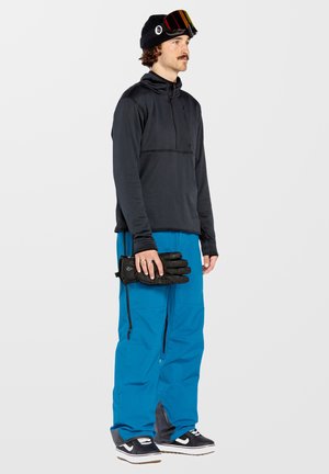 Pullover nero con mezza zip, pantaloni da neve blu e guanti neri. L'outfit presenta un tessuto testurizzato e una vestibilità strutturata.