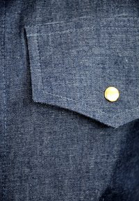 Denim w ciemnoniebieskim odcieniu, z zaostrzoną naszywaną kieszenią i złotym metalowym zatrzaskiem. Widoczne szwy dodają szczegółów teksturze.