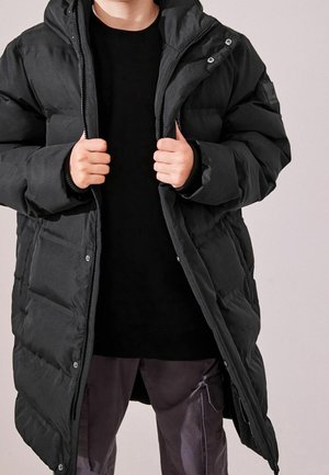 Henkilö, jolla on päällään pitkä musta toppatalvitakki mustan collegepaidan ja tummien cargo-housujen päällä, ja joka pitää takin avoimia etureunoja kiinni.