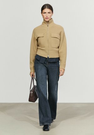 Donna con maglione a coste beige con zip, jeans scuri a gamba larga, scarpe nere, porta una piccola borsa marrone, camminando su pavimento grigio chiaro.